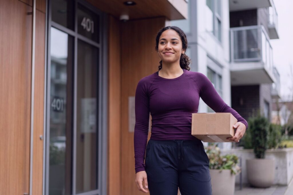 Confident content creator standing outside a modern apartment building, holding a discreet delivery box at the front door, conveying privacy, independence, and control over personal health.